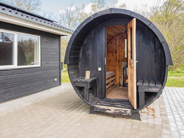 Une structure en forme de tonneau, faite de bois, avec une porte ouverte, révélant un petit espace intérieur comprenant un banc et une cheminée.