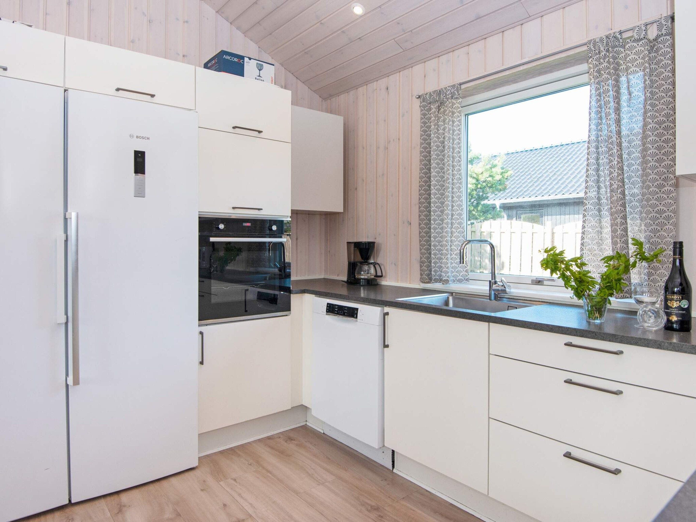 A modern kitchen with white cabinetry, a Bosch refrigerator, an oven, a coffee maker, and a window with patterned curtains.