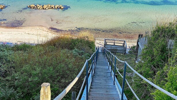 Eine Holztreppe führt zu einem Strand mit klarem Wasser und einem Felsen.