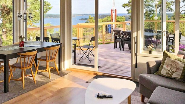 A modern living room with a view of a lake, a dining area with wooden chairs, and a large window. - 4 Star Holiday Home in Akersberga (Akersberga)
