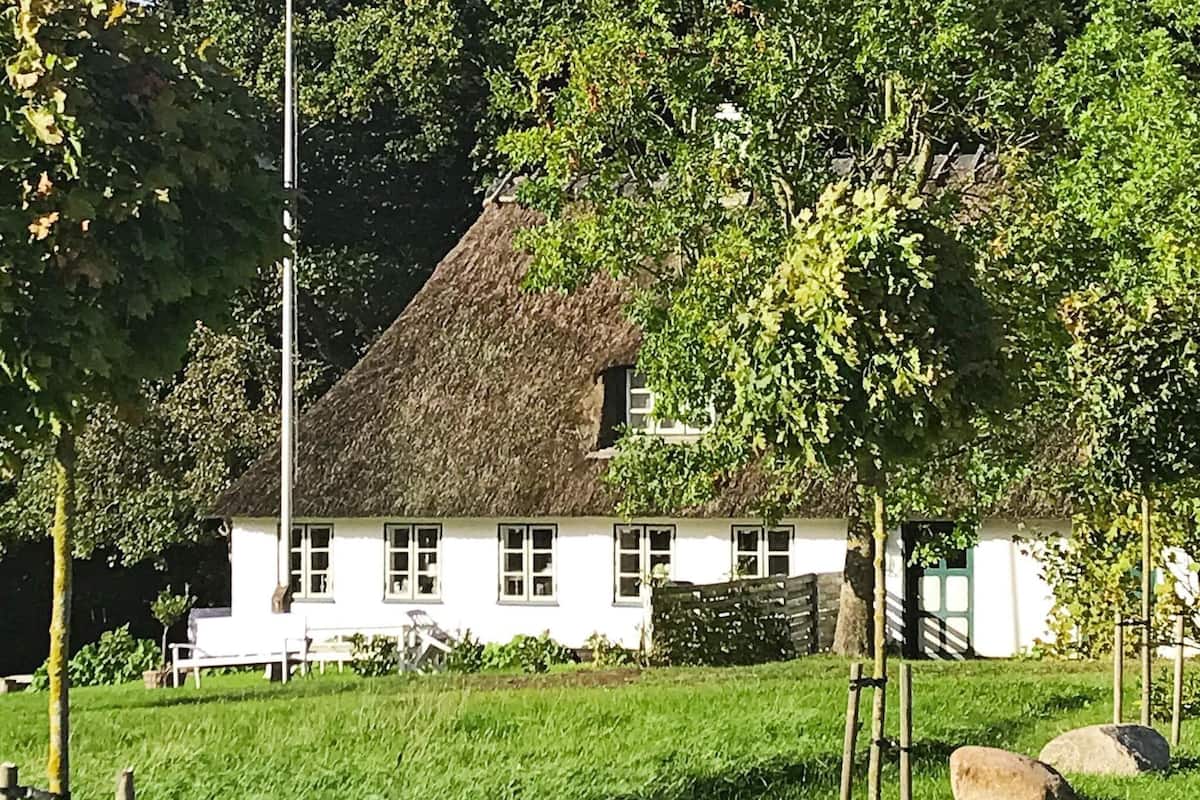 A white house with a thatched roof, surrounded by greenery and rocks.