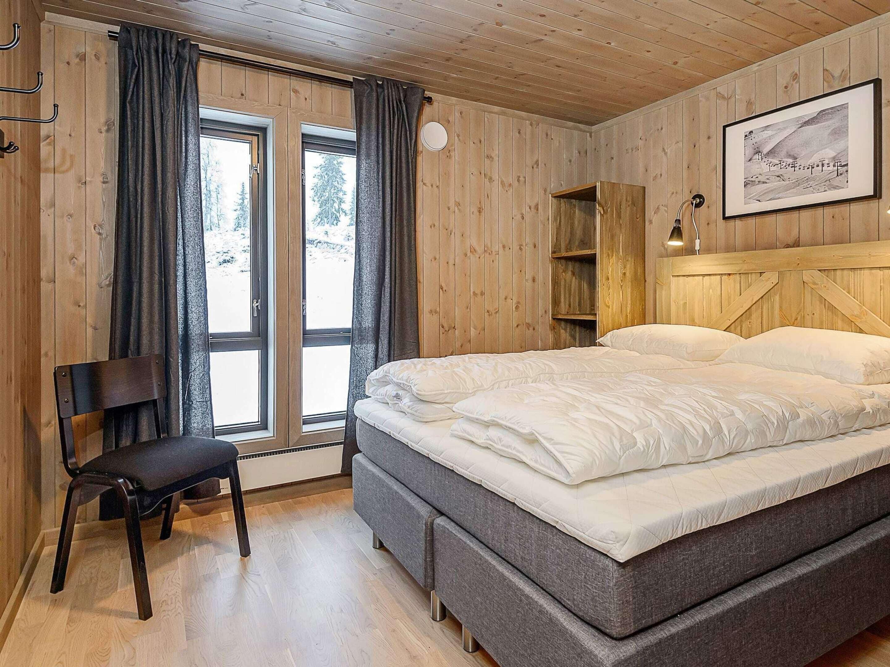 A bedroom with a bed, a chair, a wooden headboard, a window with curtains, and a framed picture on the wall.