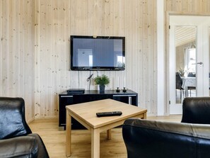 A room with a wooden paneled wall, a flat-screen TV mounted above a black entertainment unit, a wooden coffee table with a remote control, and two black leather chairs.