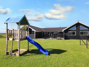 A playground with a slide and climbing structures, a covered seating area, and a building with a sloped roof and large windows.