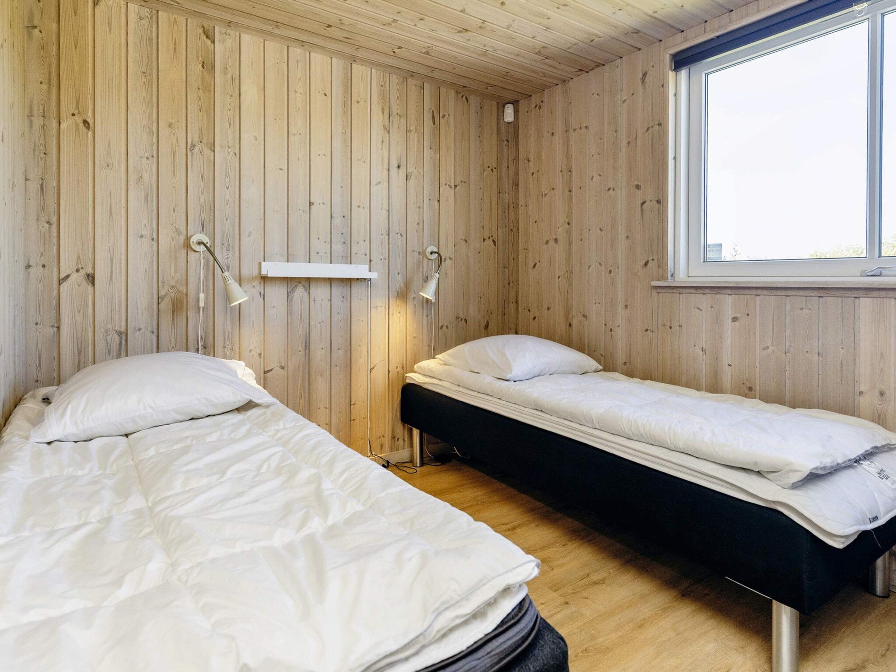 A room with two beds, wooden walls, a window, and a wall-mounted light fixture.