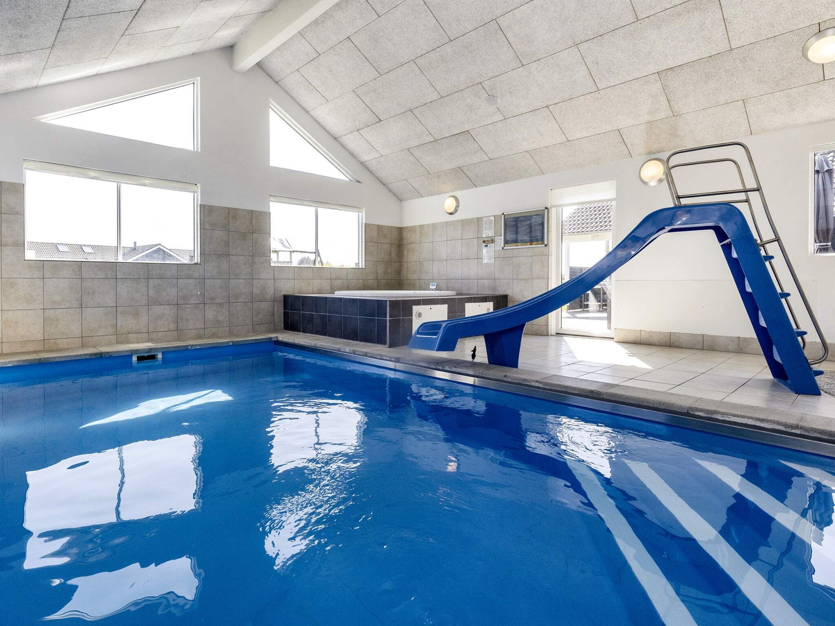 En indendørs swimmingpool med blå rutsjebane, flisebeklædte vægge og store vinduer.