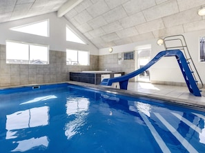 An indoor swimming pool with a blue slide, large windows, and a tiled ceiling.