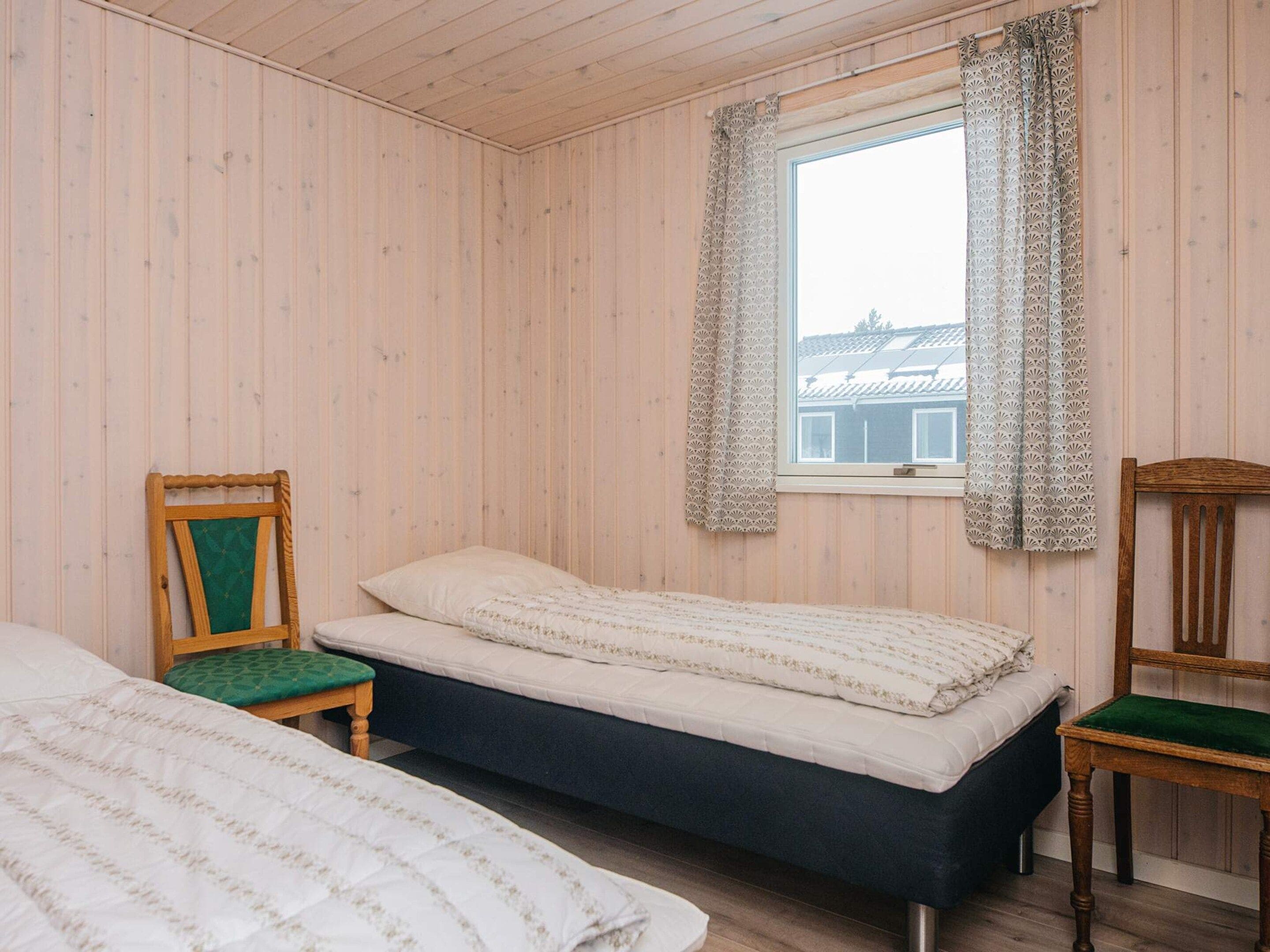 A room with two beds, a window with patterned curtains, and a chair with a green cushion.