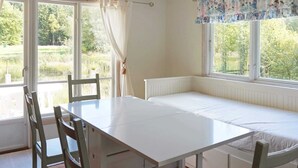 Une pièce avec une table et des bancs blancs, de grandes fenêtres avec des rideaux et une vue sur la verdure à l’extérieur.