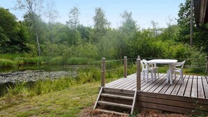 Une terrasse en bois avec une table et des chaises, donnant sur un étang bordé d’une végétation luxuriante.