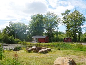 A red cabin with a porch, surrounded by a natural landscape with trees and a body of water.