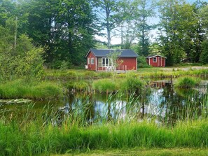 A red cabin with a porch, surrounded by a pond and dense greenery.