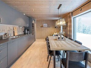 A modern kitchen with grey cabinets, a wooden dining table, and a view of snowy trees through a large window.