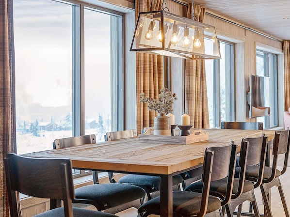 A dining area with a large wooden table, chairs, a hanging light fixture, and a view of a snowy landscape through large windows.