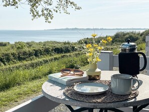A round table with a book, a pair of glasses, a vase with yellow flowers, a coffee pot, and a mug on a woven placemat, overlooking a grassy landscape and the sea. - 5 Star Holiday Home in Aabenraa (Aabenraa)