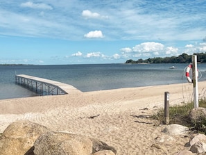 A beach with a wooden pier extending into calm waters, a lifebuoy on a post, and a distant shoreline with trees.