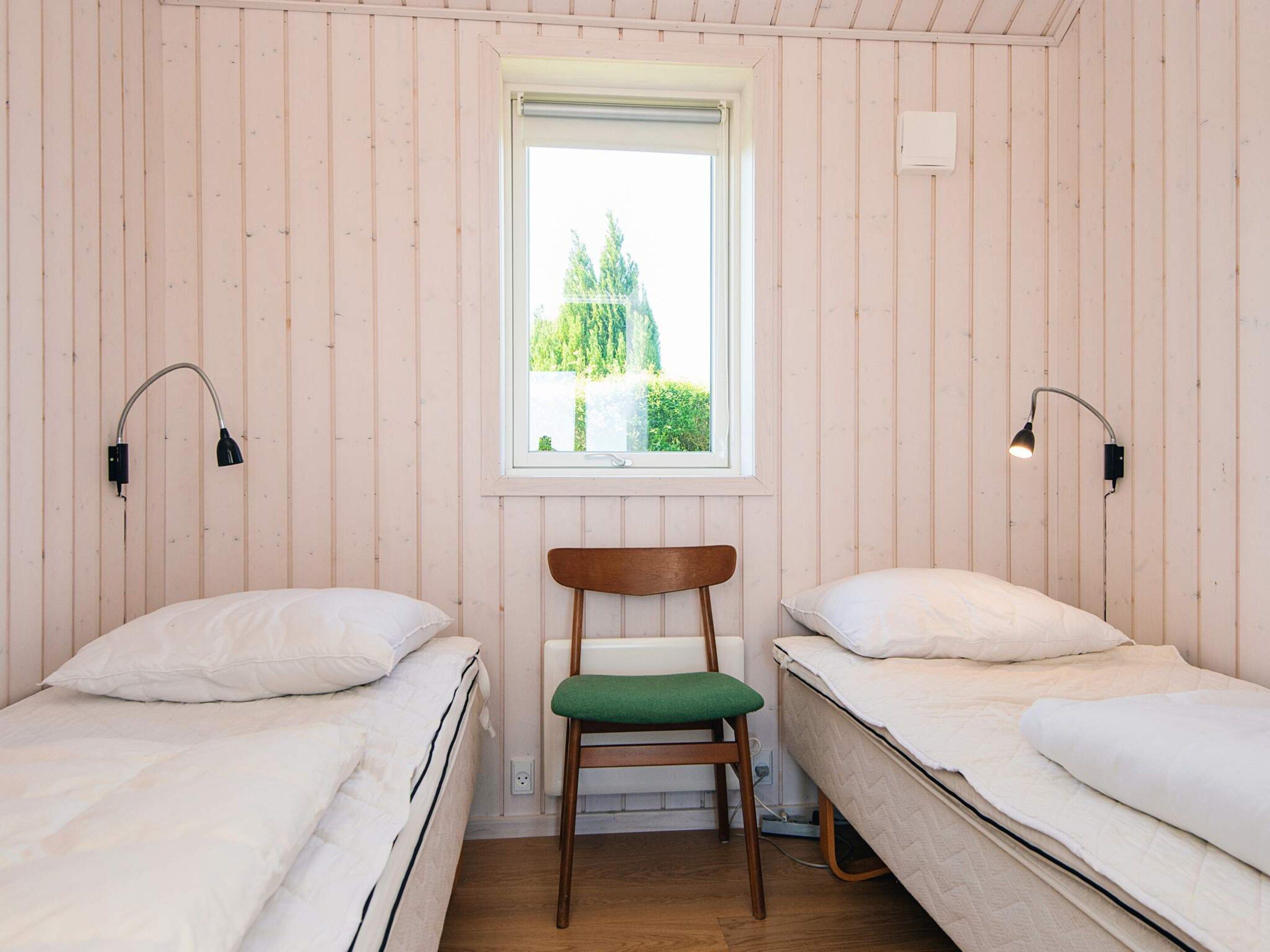 A room with two single beds, a wooden chair, a window with a view of greenery, and two wall-mounted lamps.