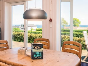 Uma sala de jantar com mesa, cadeiras, um livro e vista para o mar através de grandes janelas.
