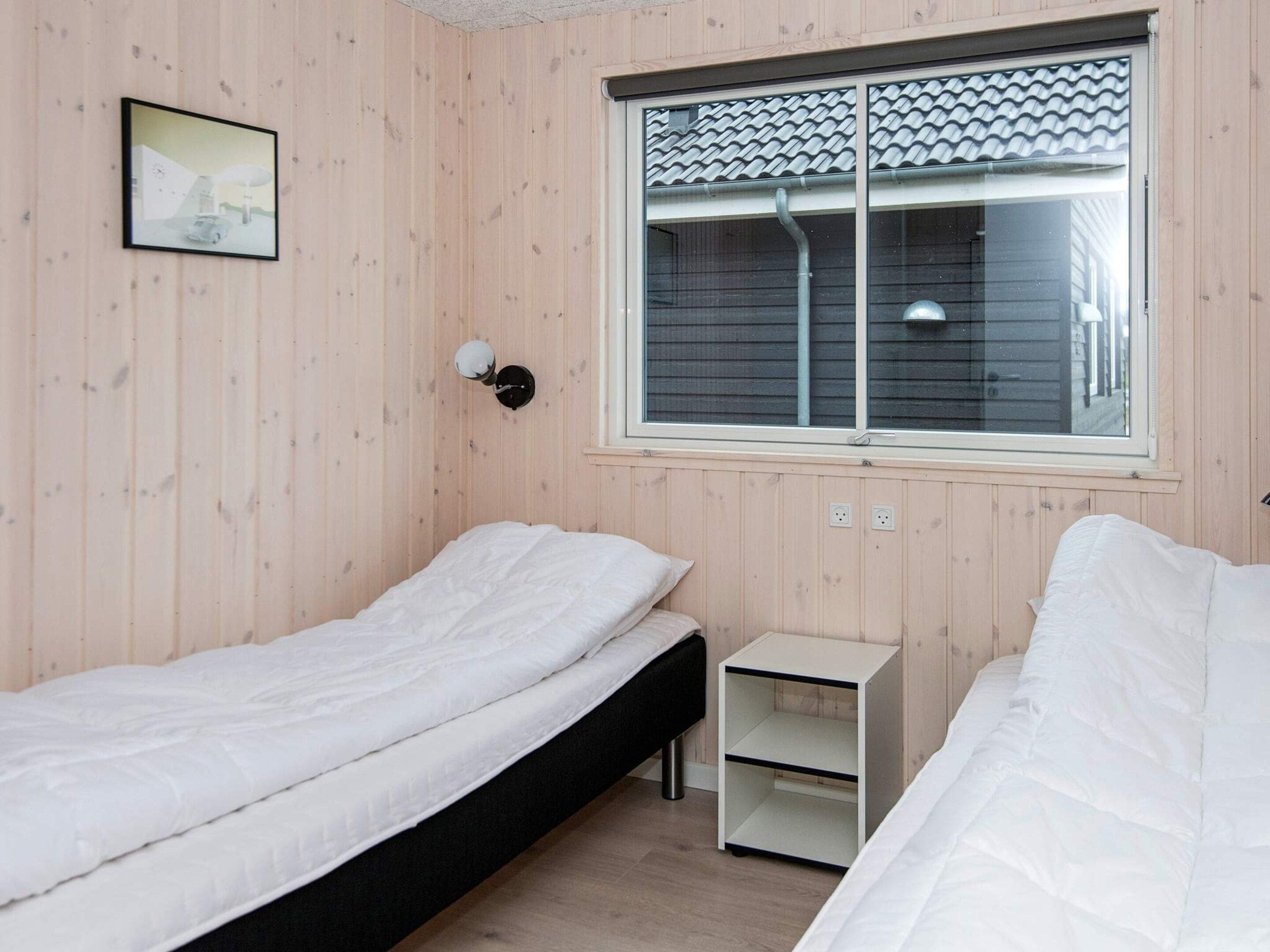 A room with two single beds, a small bedside table, a wall-mounted lamp, and a window with a view of a building.