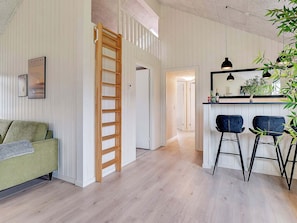 A room with a wooden ladder, a green sofa, two bar stools, a bar counter with bottles, and a hanging plant. - 5 Star Holiday Home in Norre Nebel (Norre Nebel)