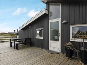 A wooden deck with a bench and a table, a door with a window, and potted plants. - 5 Star Holiday Home in Ringkobing (Ringkobing)
