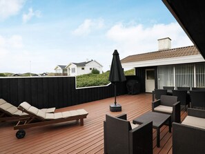 Une terrasse avec des chaises longues et une table, un barbecue abrité, et une vue sur les maisons au loin.