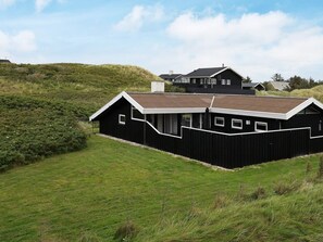 Un bâtiment noir avec un toit en pente, entouré d’un champ herbeux et d’une colline.