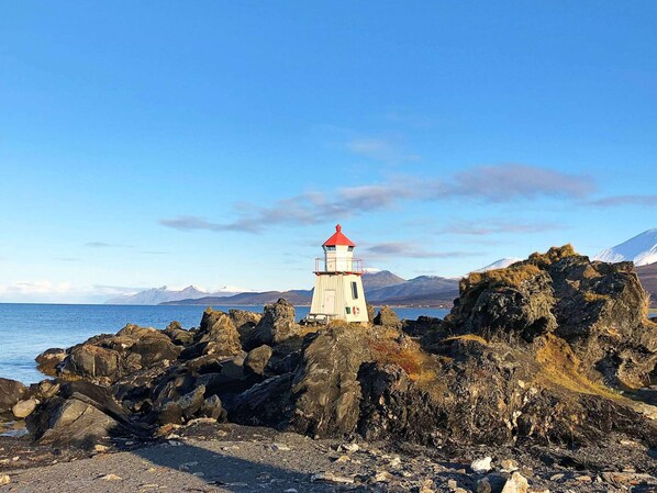 Un phare se dresse sur un terrain rocheux, sous un ciel dégagé, avec des montagnes qui se profilent à l’horizon.