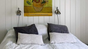 A bed with white linens and two dark pillows, a painting of sunflowers above it, and two wall-mounted lamps.