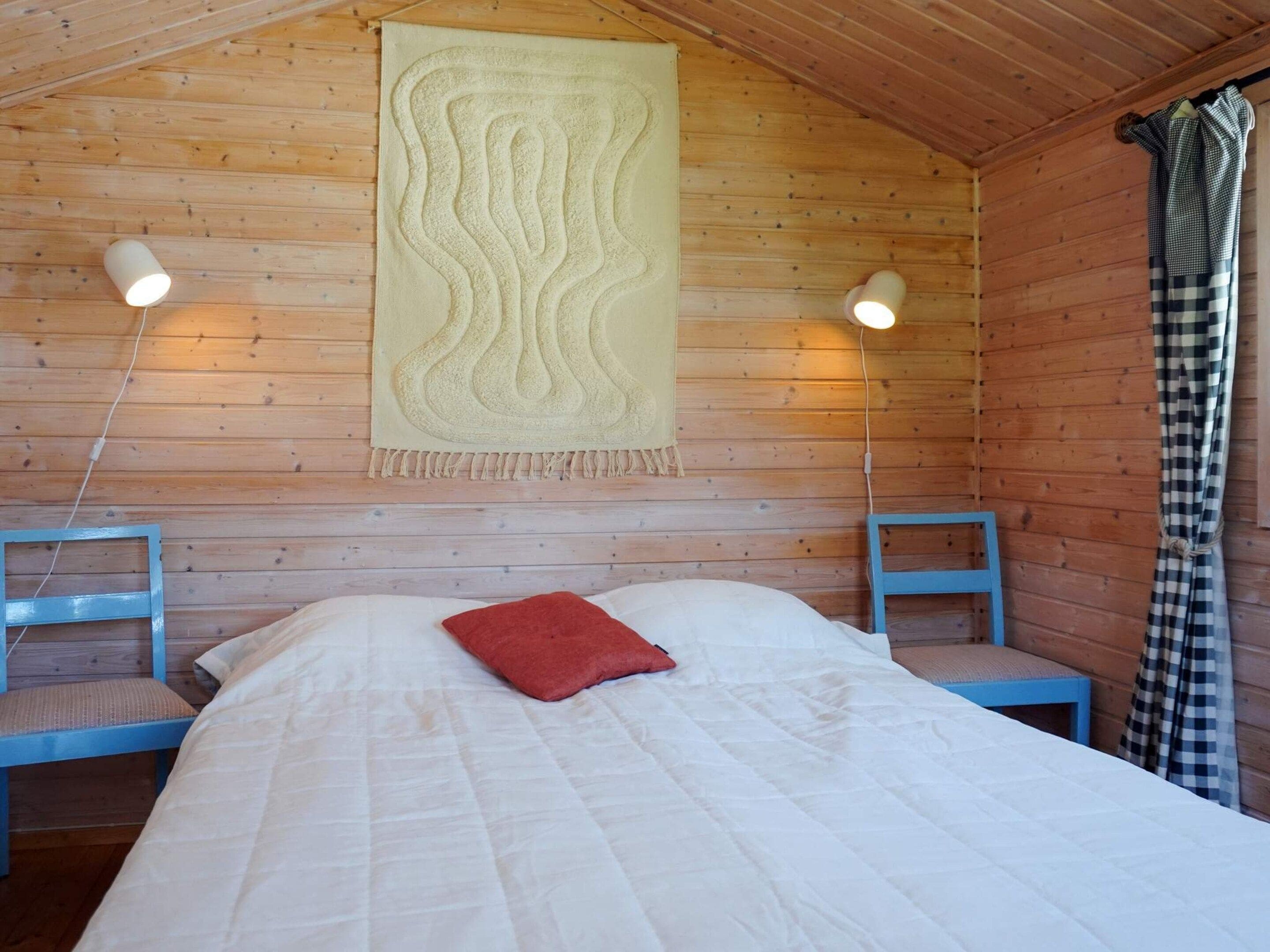 Una habitación de cabaña de madera con cama, dos sillas, un tapiz colgante y una ventana con cortinas.