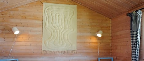 A wooden cabin room with a bed, two chairs, a hanging tapestry, and a window with curtains.