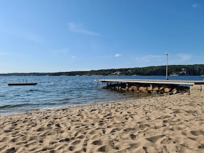 Una playa arenosa con un muelle que se adentra en aguas tranquilas, un bote pequeño y una colina boscosa al fondo.