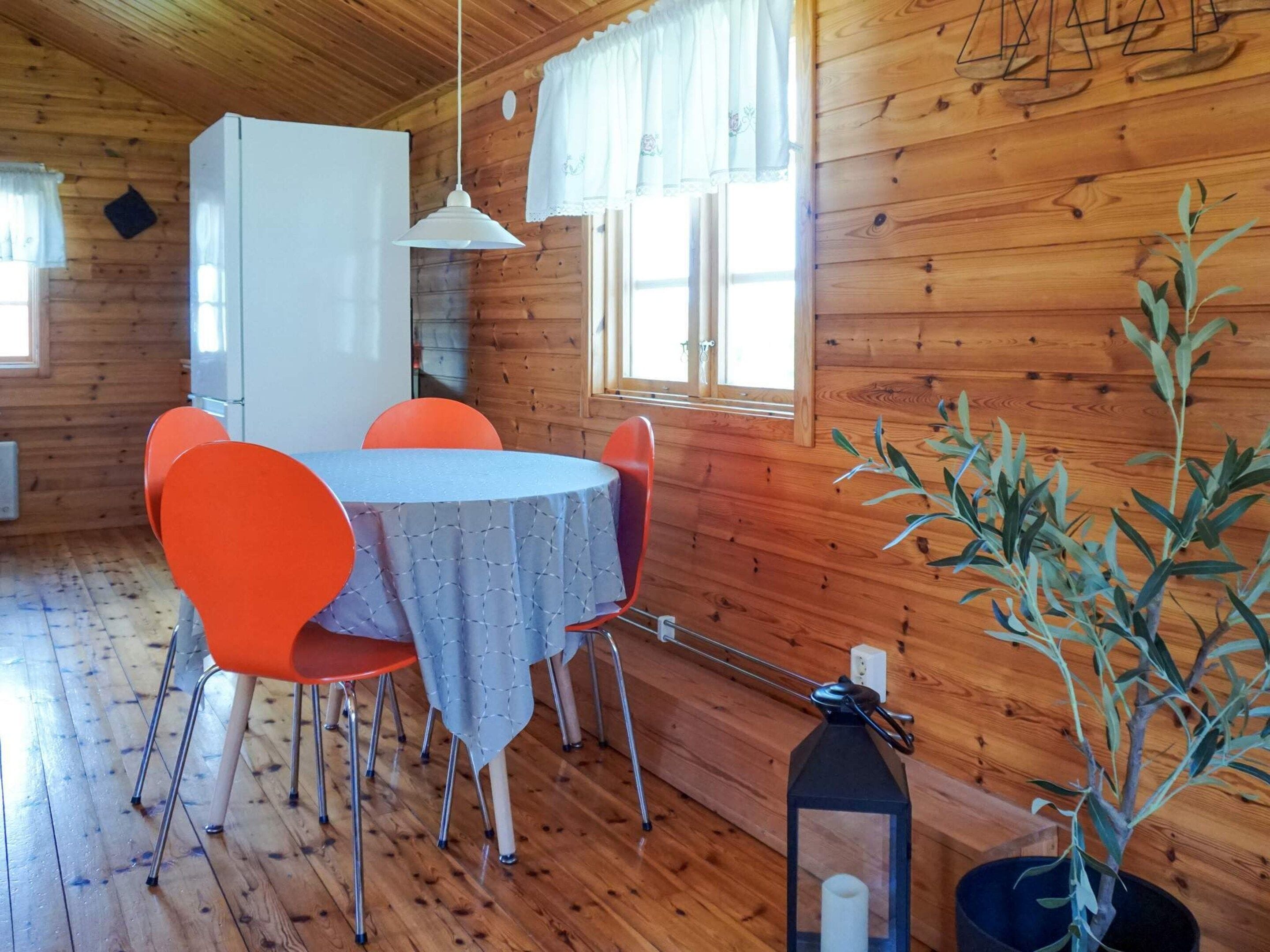 Interior de una cabaña de madera con una mesa de comedor, sillas anaranjadas, un refrigerador blanco y una planta en maceta.