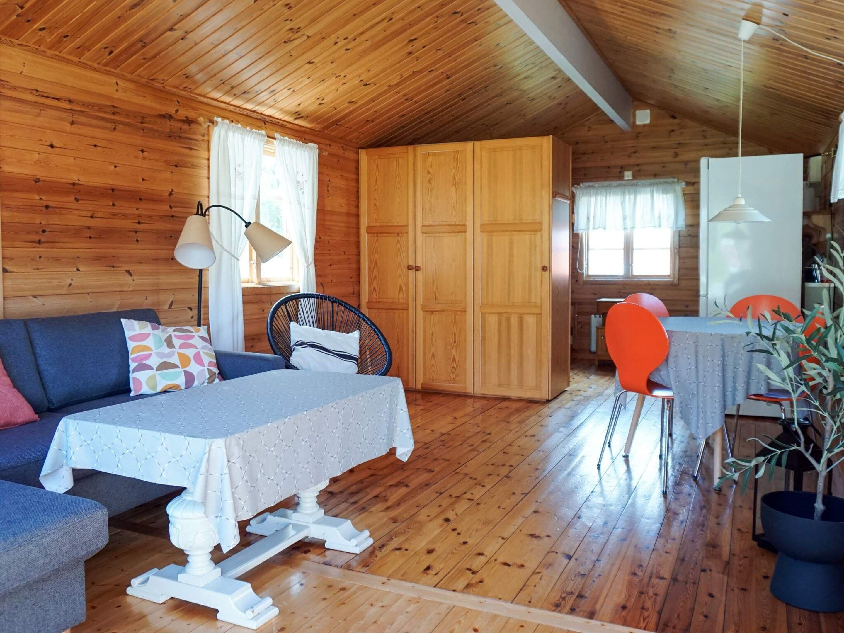 A wooden cabin interior with a sofa, dining table, chairs, a lamp, a wardrobe, and a potted plant.