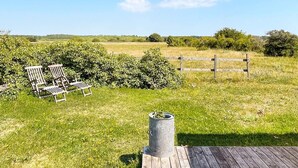 A wooden deck with a metal trash can, a wooden fence, and a grassy field with a bench and chairs in the foreground. - 4 Person Holiday Home in Gotlands.tofta (Gotlands Tofta)