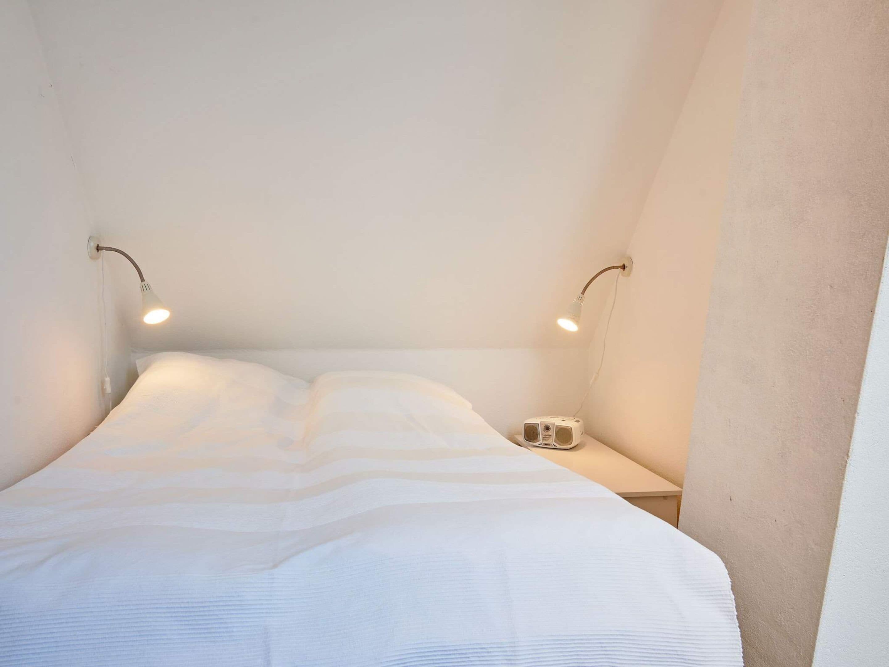 Ein kleines Zimmer mit einem Einzelbett, einer weißen Bettdecke, zwei Nachttischlampen und einem kleinen Nachttisch mit einem Radio.