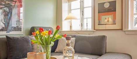 A living room with a grey sofa, a round wooden coffee table with a vase of flowers, candles, and glasses, and a window with a lamp.