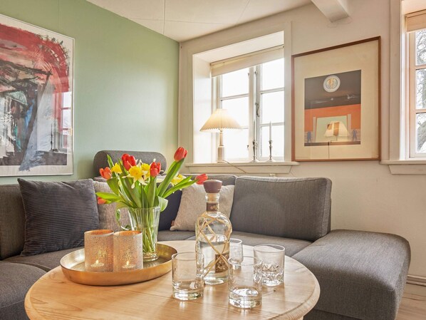 A living room with a grey sofa, a round wooden coffee table with a vase of flowers, candles, and glasses, and a window with a lamp.