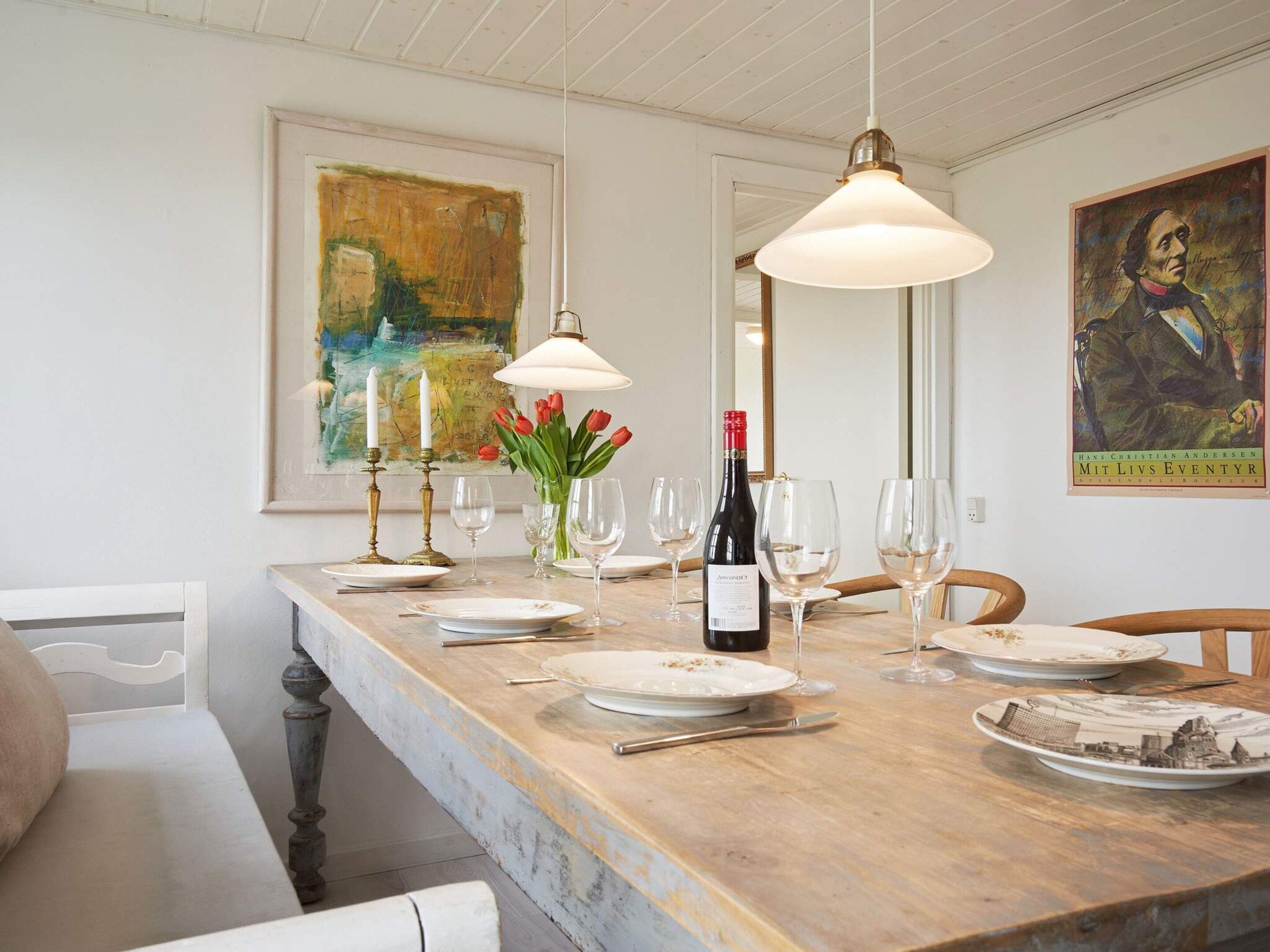 A dining table set with plates, glasses, and a bottle of wine, with artwork on the walls and a pendant light above.