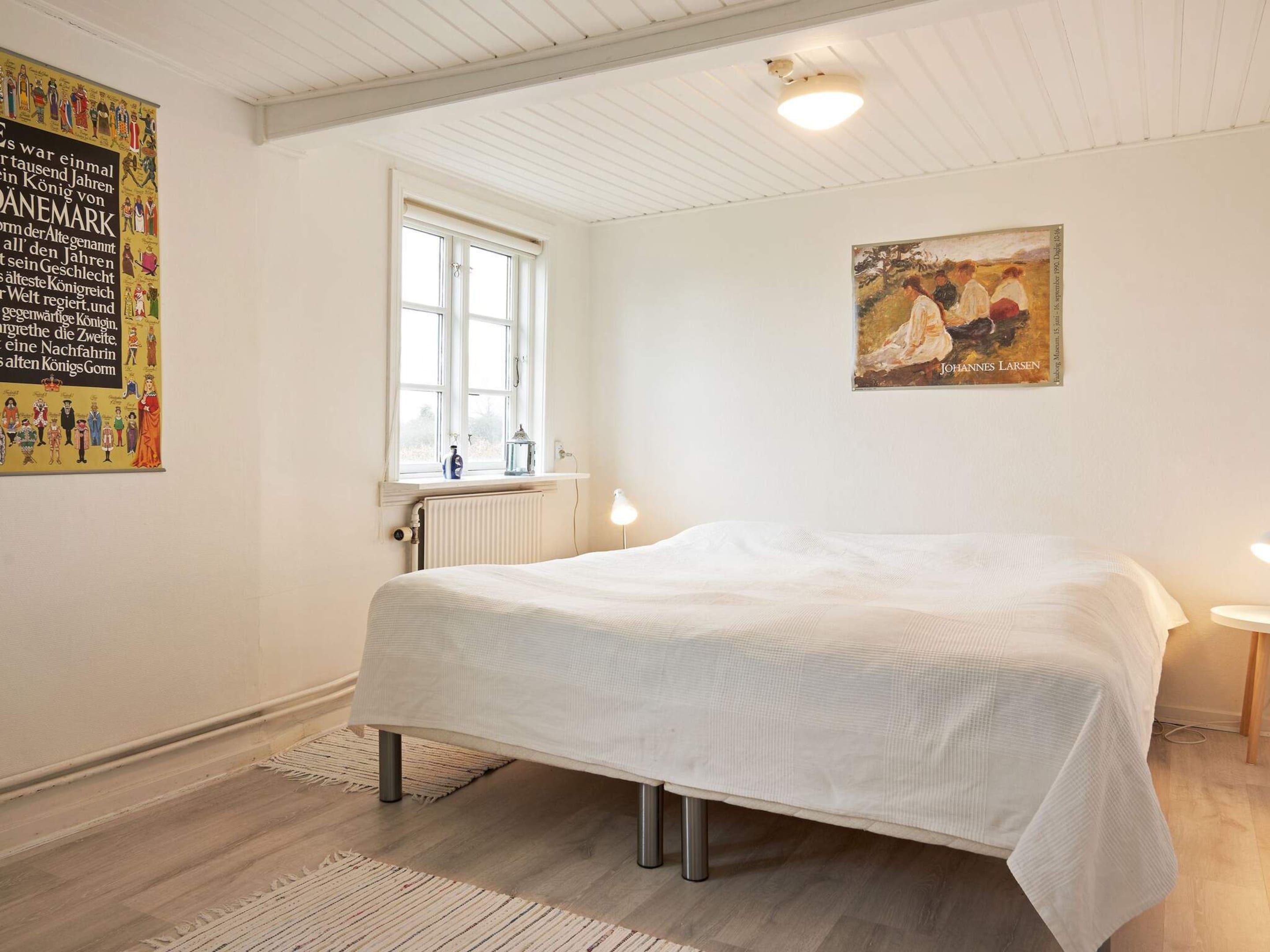A bedroom with a bed, a window, a radiator, a painting, and a poster on the wall.