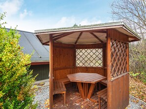 A wooden outdoor seating area with a table and benches, a lattice window, and a covered roof structure.