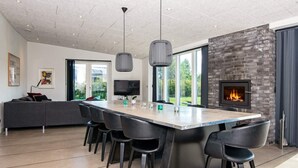 A modern kitchen with a large island, black bar stools, a fireplace, and a large window overlooking a garden. - Luxury Lakeside Retreat With Spa-by Traum (Glesborg)