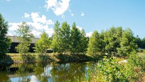 A serene pond surrounded by lush greenery and trees, with a building in the background. - Luxury Lakeside Retreat With Spa-by Traum (Glesborg)