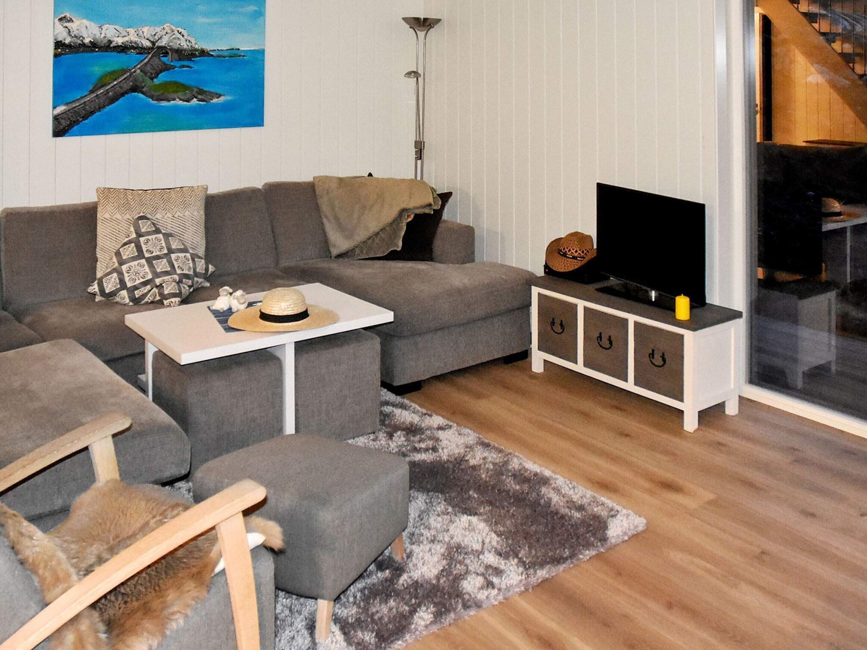 Ein modernes Wohnzimmer mit einem grauen Sofa, einem kleinen weißen Tisch, einer TV-Konsole mit Fernseher und einem Gemälde mit Küstenlandschaft.