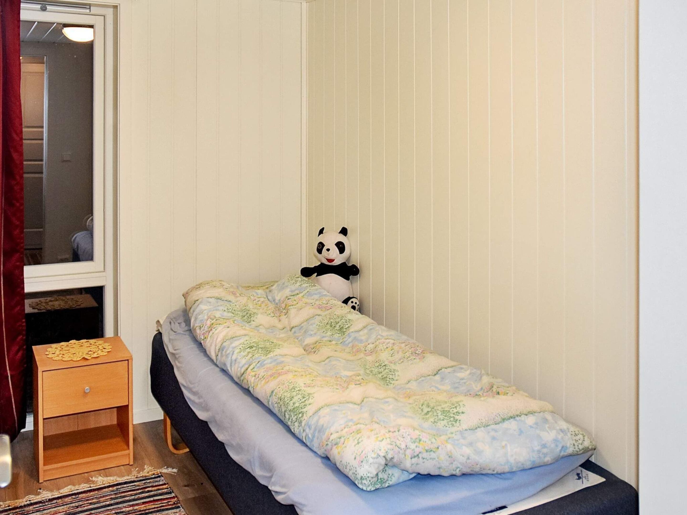 A small, single-bed room with a window, a wooden nightstand, and a panda plush toy on the bed.