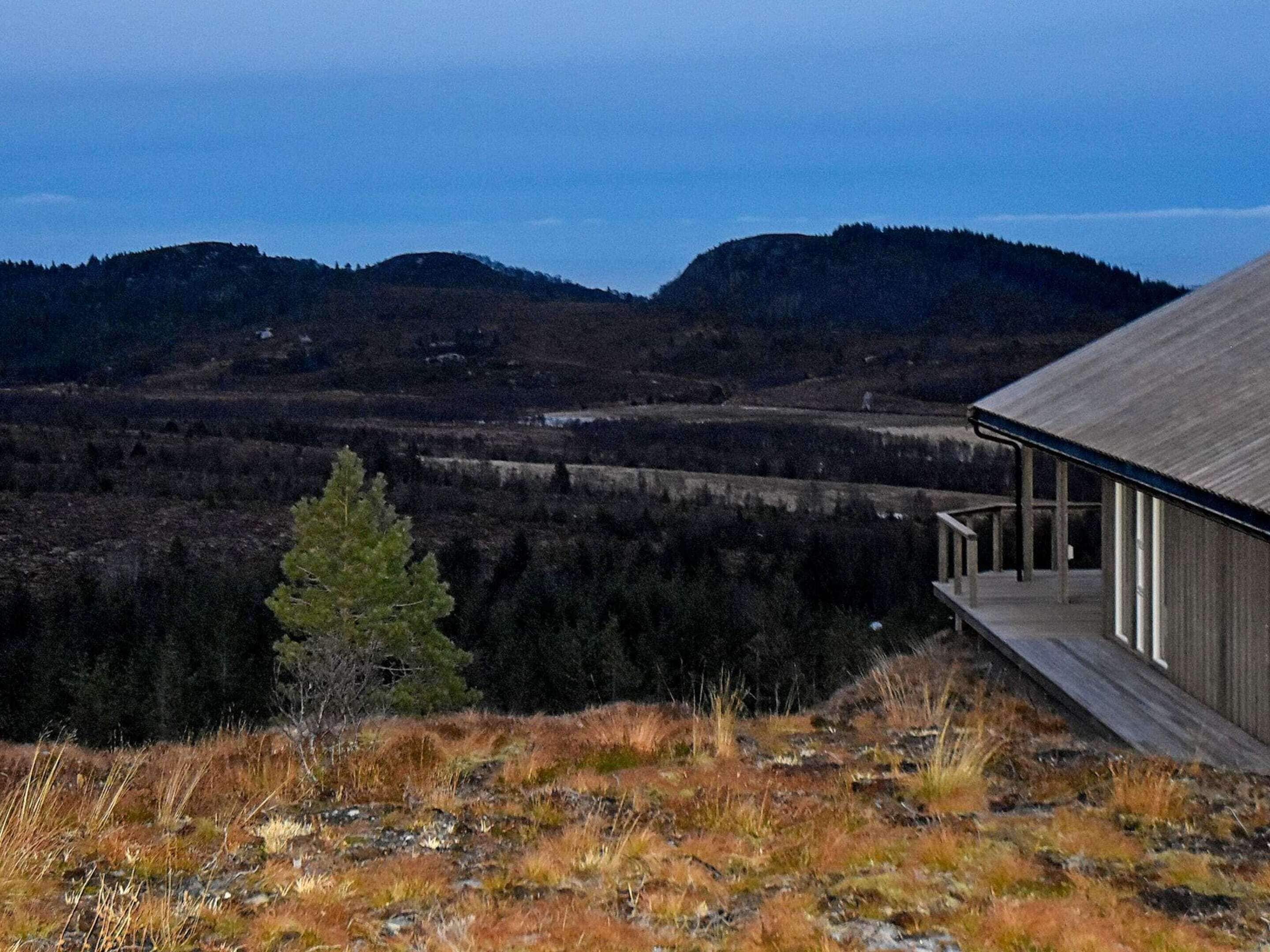 A modern building with a flat roof and a covered porch, surrounded by a hilly landscape with dense forests.