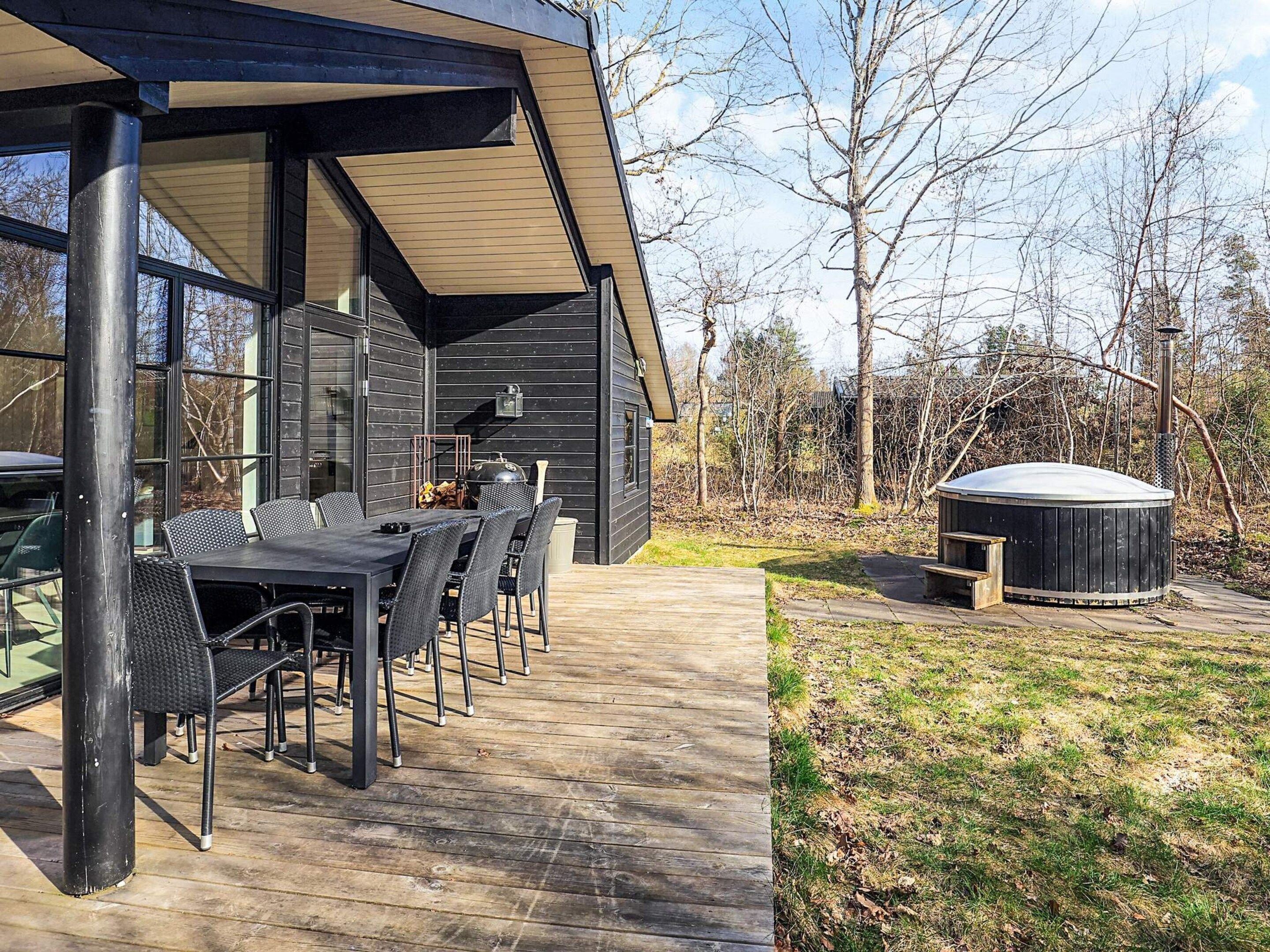 A modern outdoor dining area with a long table and chairs, a barbecue grill, and a hot tub in a wooded setting.