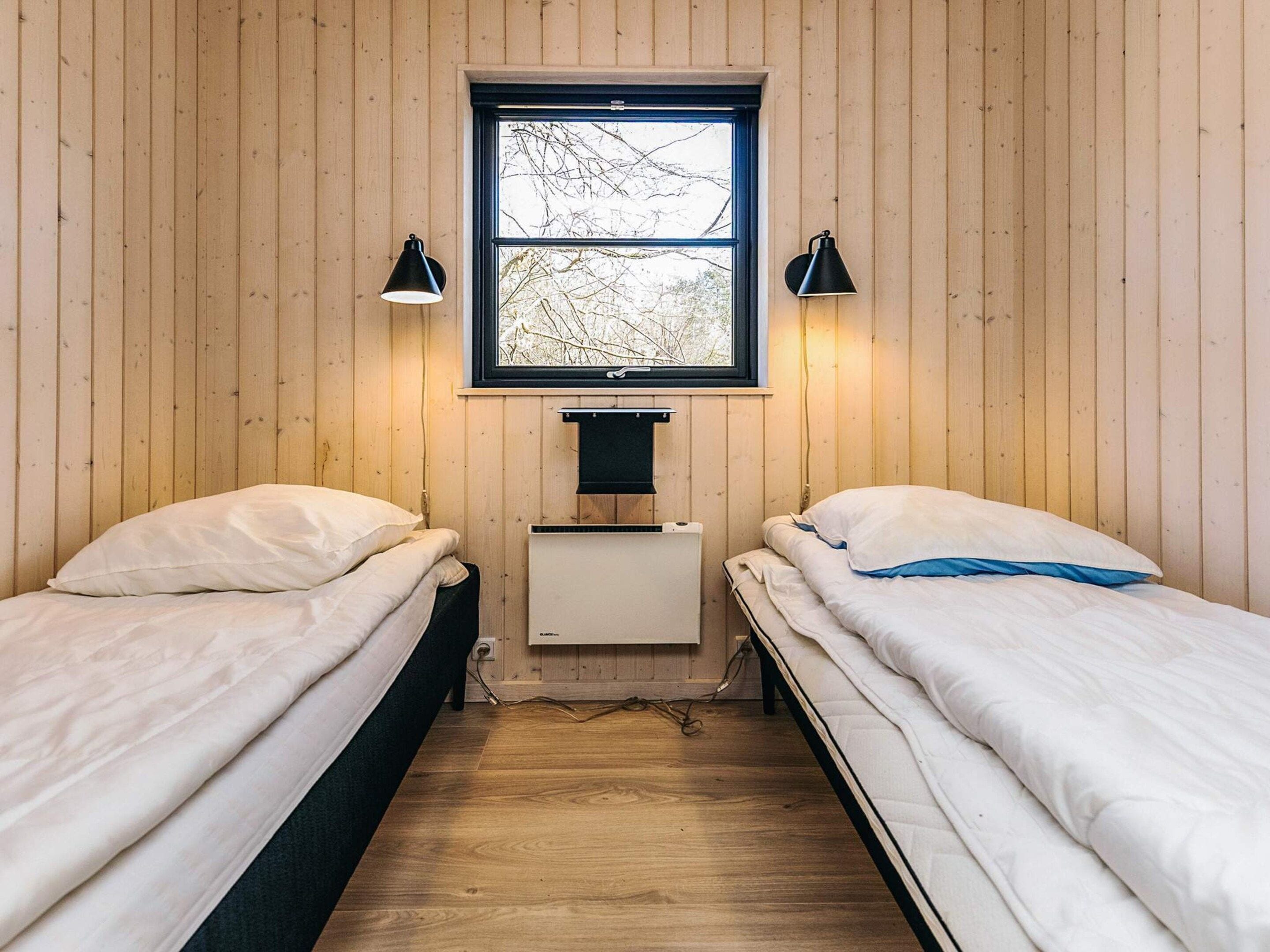 Una habitación pequeña y acogedora con dos camas individuales, una ventana que da a árboles y un escritorio pequeño con una lámpara.