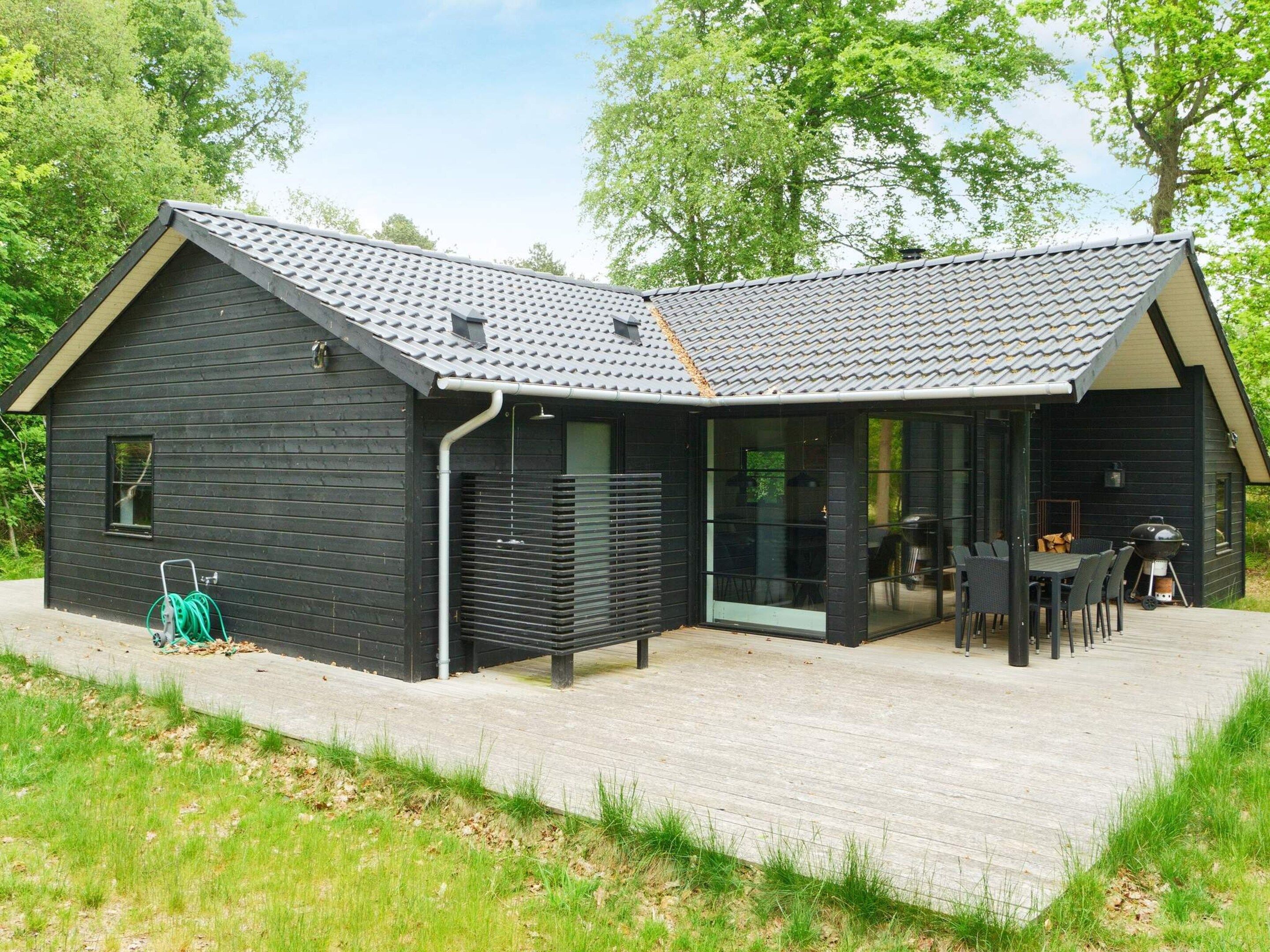 A modern black building with a tiled roof, a covered patio with outdoor furniture, and a hose reel on the ground.