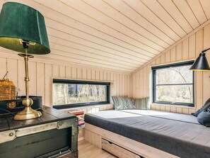 Een knus houten huisje van binnen, met een bed, twee ramen, een lamp en een kleine tafel.